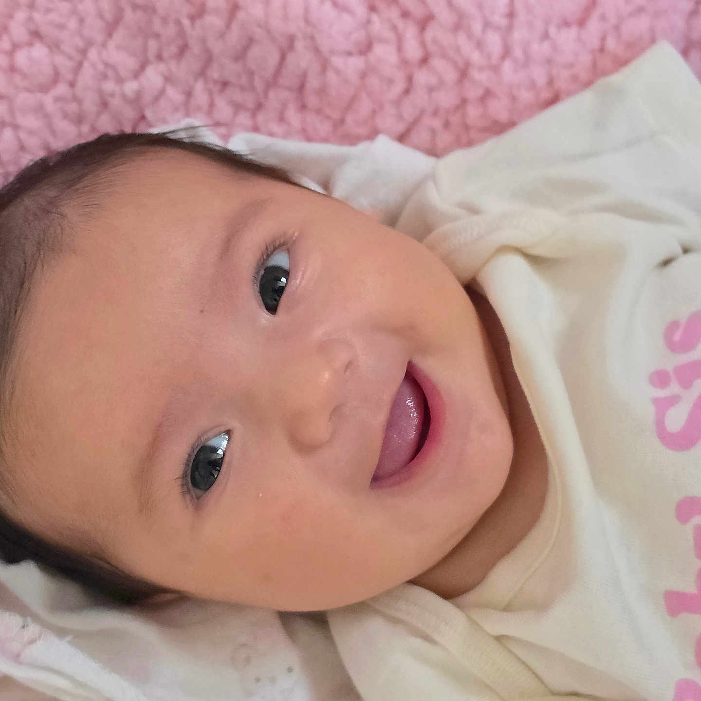 Klaritie is registered to the contest to win money with this photo: baby, infant, child, face, portrait, smiling, happy, lying_down, blanket, pink, white_clothing, onesie, indoor, close_up, brown_hair, eyes, mouth, cozy, textile, photography