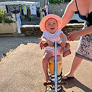 Maya a rejoint le concours — aidez-le/la à gagner de superbes lots ! toddler, child, spring_rocker, hat, smile, outdoor, sunlight, playground, adult, supporting_hand, clothing, market, pavement, stone_wall, greenery, footwear, summer, happy, person, toy