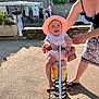 toddler, child, spring_rocker, hat, smile, outdoor, sunlight, playground, adult, supporting_hand, clothing, market, pavement, stone_wall, greenery, footwear, summer, happy, person, toy
