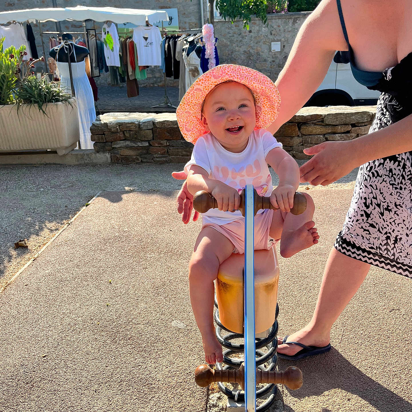 Maya a rejoint le concours — aidez-le/la à gagner de superbes lots ! adult, child, clothing, footwear, greenery, happy, hat, market, outdoor, pavement, person, playground, smile, spring_rocker, stone_wall, summer, sunlight, supporting_hand, toddler, toy
