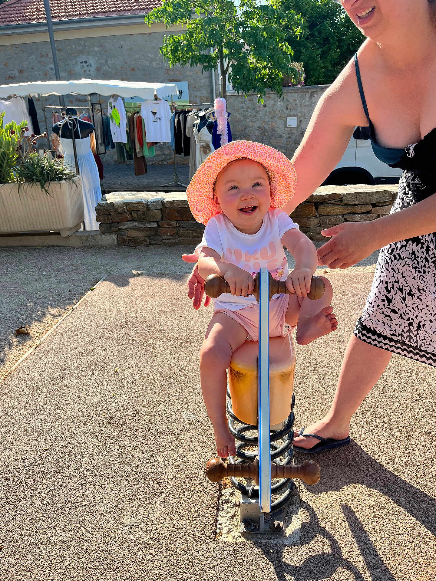 Maya a rejoint le concours — aidez-le/la à gagner de superbes lots ! toddler, child, spring_rocker, hat, smile, outdoor, sunlight, playground, adult, supporting_hand, clothing, market, pavement, stone_wall, greenery, footwear, summer, happy, person, toy