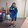 Beckett is registered to the contest to win money with this photo: child, toddler, smiling, portrait, standing, indoors, blue_coat, winter_clothing, hood, colorful_slippers, superhero_pose, playful, door, carpet, chair, scooter, boxes, cluttered_room, household_items, happy