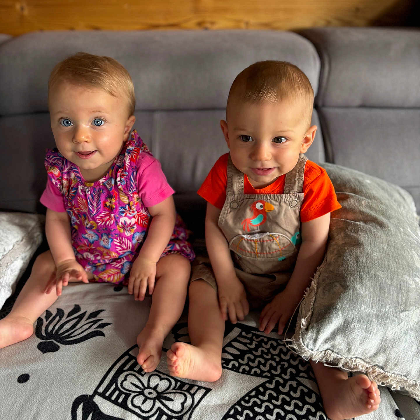 Mia a rejoint le concours — aidez-le/la à gagner de superbes lots ! baby, blanket, child, couch, curious, dress, face, feet, furniture, indoor, orange_shirt, overalls, pattern, pillow, pink_shirt, sitting, smiling, toddler, two_children, wooden_wall
