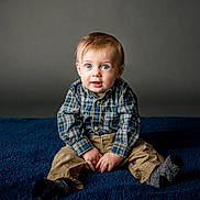 Alfred is registered to the contest to win money with this photo: baby, child, plaid_shirt, khaki_pants, socks, blue_blanket, gray_background, sitting, portrait, indoor, curious_expression, hands, face, eyes, young_child, person, clothing, studio, cute, photography