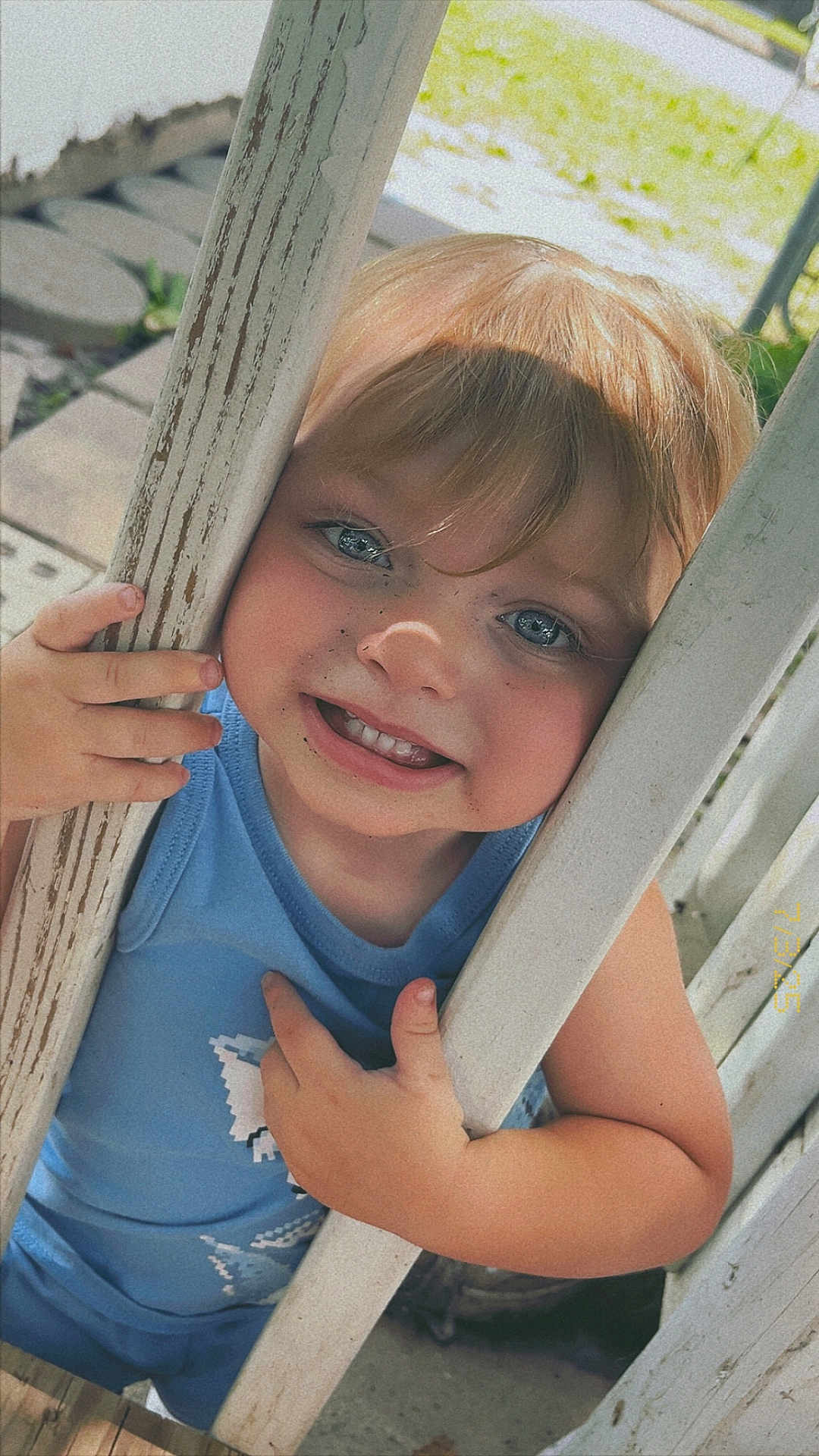 Kingston is registered to the contest to win money with this photo: child, toddler, smiling, blue_shirt, outdoor, wooden_bars, face, hands, sunlight, grass, playful, freckles, blonde_hair, portrait, close_up, happy, person, summer, daylight, cute