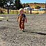 child, blonde_hair, overalls, striped_shirt, gravel, road, rural, fence, trees, house, road_sign, sky, clouds, outdoor, daylight, footwear, person, young_child, nature, fence_post