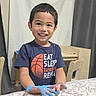 child, boy, smiling, paint, blue_paint, table, tablecloth, indoor, curtain, furniture, shirt, basketball_graphic, happy, hands, young, portrait, casual_clothing, wooden_chair, home, playful