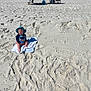 baby, child, towel, beach, sand, sun_hat, footprints, writing_in_sand, swimwear, outdoor, sunny, summer, vacation, infant, sitting, blue_shirt, colorful_hat, barefoot, chair, bag
