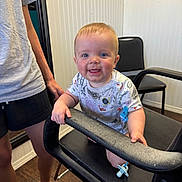 Oliver is registered to the contest to win money with this photo: baby, child, smiling, indoor, chair, black_chair, person, hand, clothing, pacifier, floor, wood_floor, wall, white_wall, shorts, sandals, adult, happy, face, blue_eyes