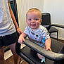 baby, child, smiling, indoor, chair, black_chair, person, hand, clothing, pacifier, floor, wood_floor, wall, white_wall, shorts, sandals, adult, happy, face, blue_eyes