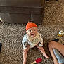baby, orange_hat, bib, carpet, sitting, smiling, diaper, spatula, couch, floor, person, sock, leg, toy, indoor, child, happy, home, clothing, furniture