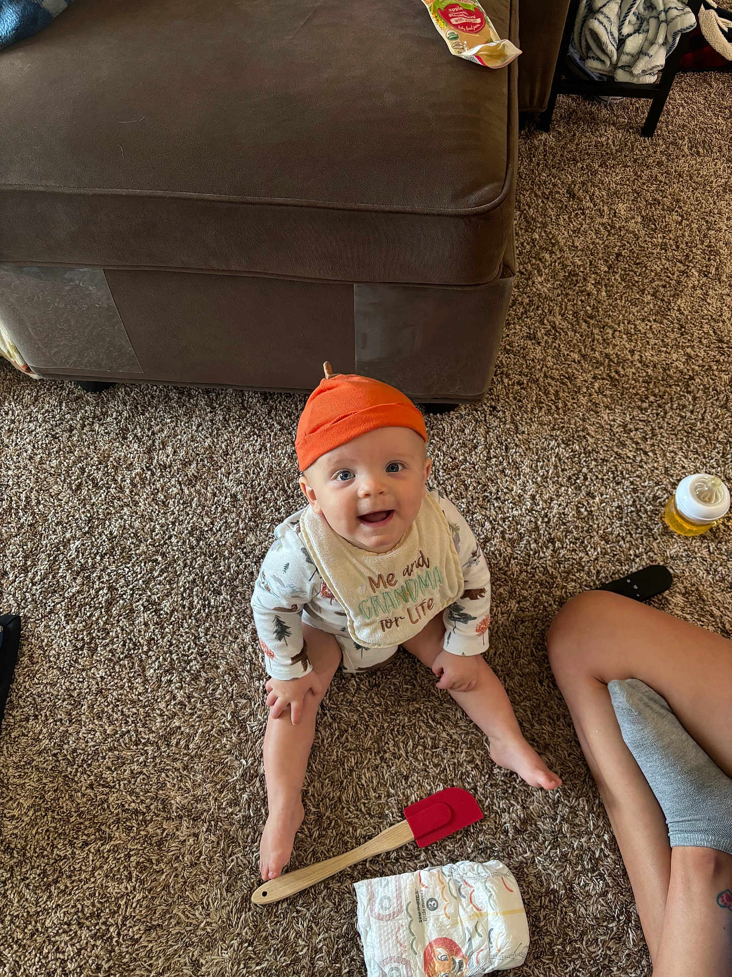 Oliver is registered to the contest to win money with this photo: baby, orange_hat, bib, carpet, sitting, smiling, diaper, spatula, couch, floor, person, sock, leg, toy, indoor, child, happy, home, clothing, furniture