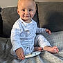 baby, child, pajamas, white_clothing, remote_control, couch, gray_couch, smile, sitting, indoor, face, happy, foot, hand, furniture, child_playing, home, infant, person, cozy