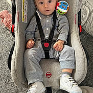 Diego is registered to the contest to win money with this photo: baby, child, car_seat, toy, sneakers, grey_clothing, indoor, carpet, safety_harness, curious, cute, infant, footwear, play, colorful_toy, seat, person, young_child, looking_at_camera, casual_clothing