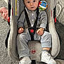baby, child, car_seat, toy, sneakers, grey_clothing, indoor, carpet, safety_harness, curious, cute, infant, footwear, play, colorful_toy, seat, person, young_child, looking_at_camera, casual_clothing