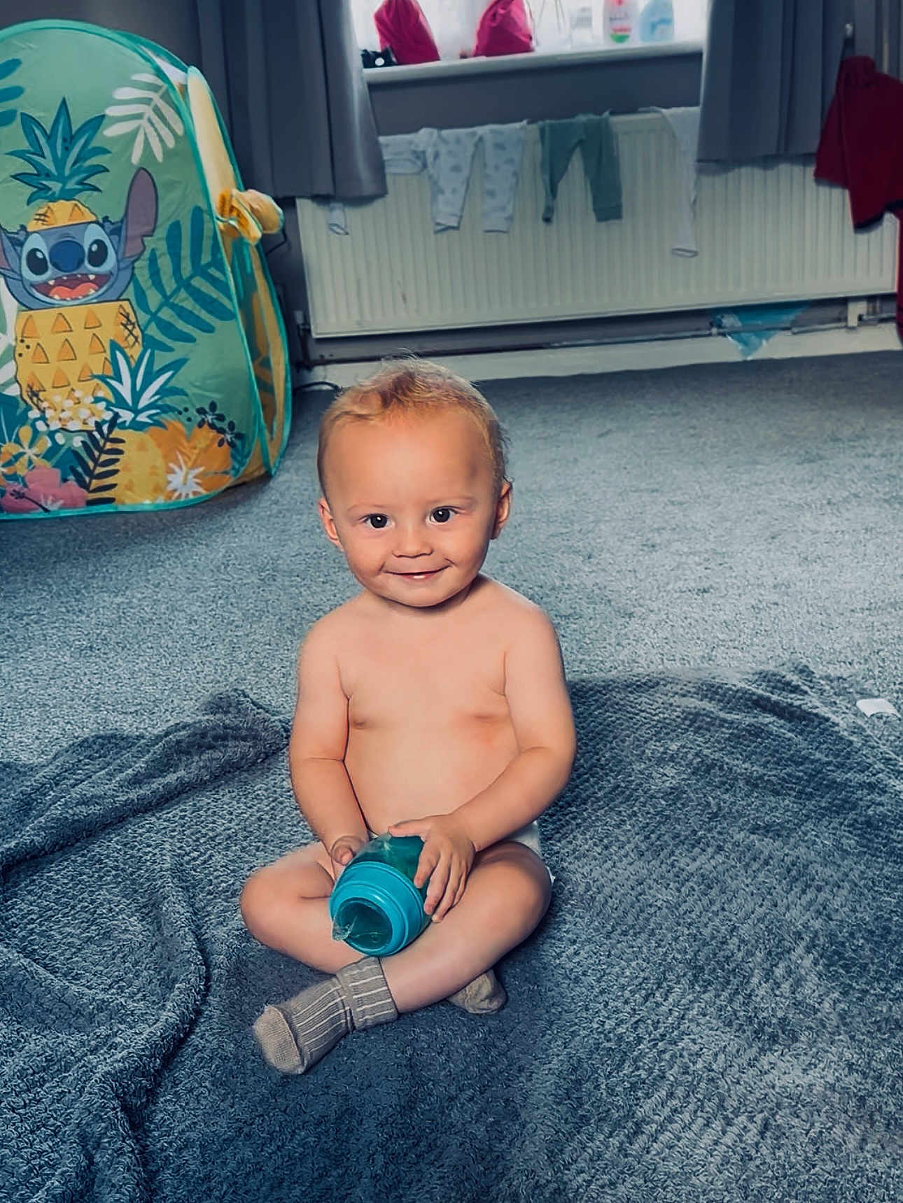 Diego is registered to the contest to win money with this photo: baby, child, smile, sitting, blanket, sippy_cup, indoor, carpet, toy, laundry, window, curtains, sock, playtime, happy, home, radiator, clothing, floor, person