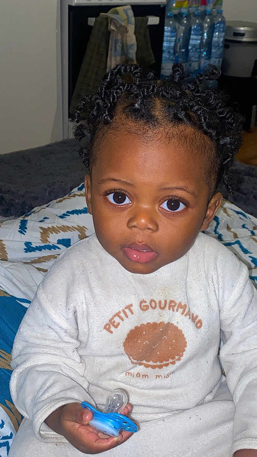 Kaecy participe au concours pour gagner de l'argent avec cette photo : child, toddler, baby, pacifier, pajamas, onesie, curly_hair, big_eyes, portrait, closeup, indoor, bed, blanket, hand, face, expression, mouth, bedding, water_bottles, room