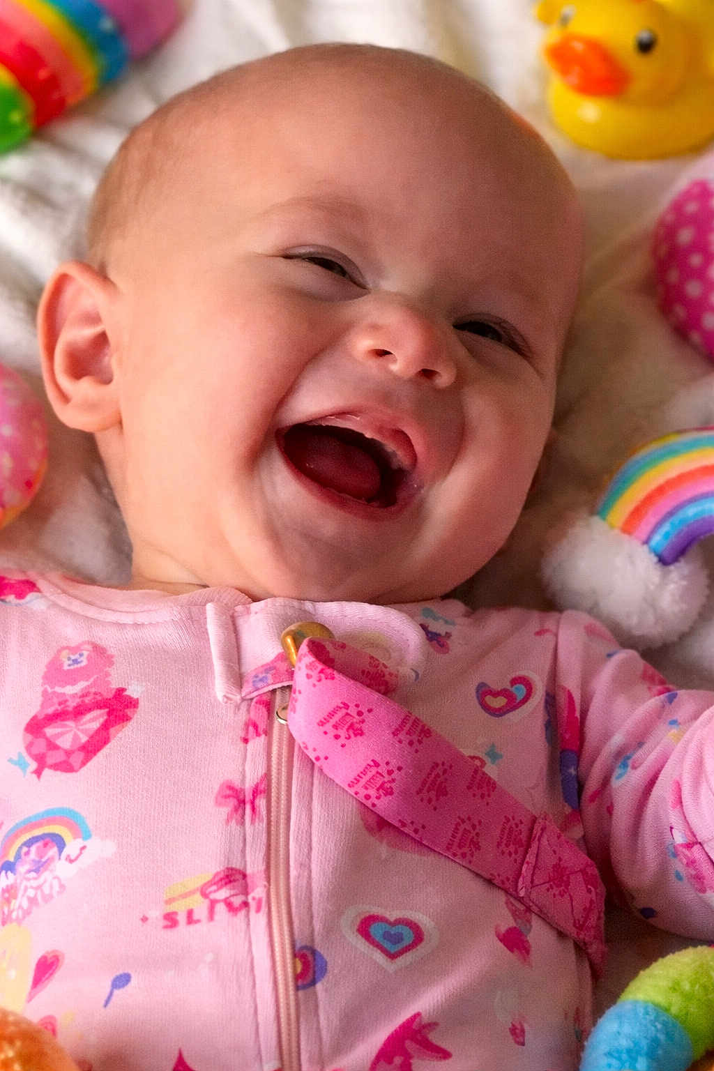 Paizlee is registered to the contest to win money with this photo: baby, infant, smile, laughing, face, onesie, pink_clothing, rubber_duck, rainbow_toy, plush_toy, close_up, portrait, mouth_open, eyes_squinting, cheek, bedding, toy, pacifier_clip, cute, colorful