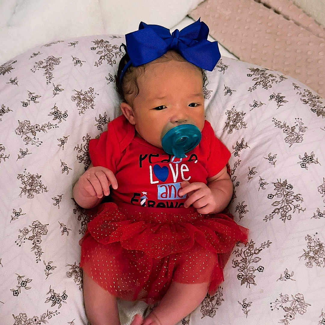 China is registered to the contest to win money with this photo: baby, bedroom, blanket, blue_bow, child, cozy, cute, floral_pattern, hands, headband, indoors, infant, newborn, pacifier, pillow, portrait, red_outfit, resting, small_feet, tutu