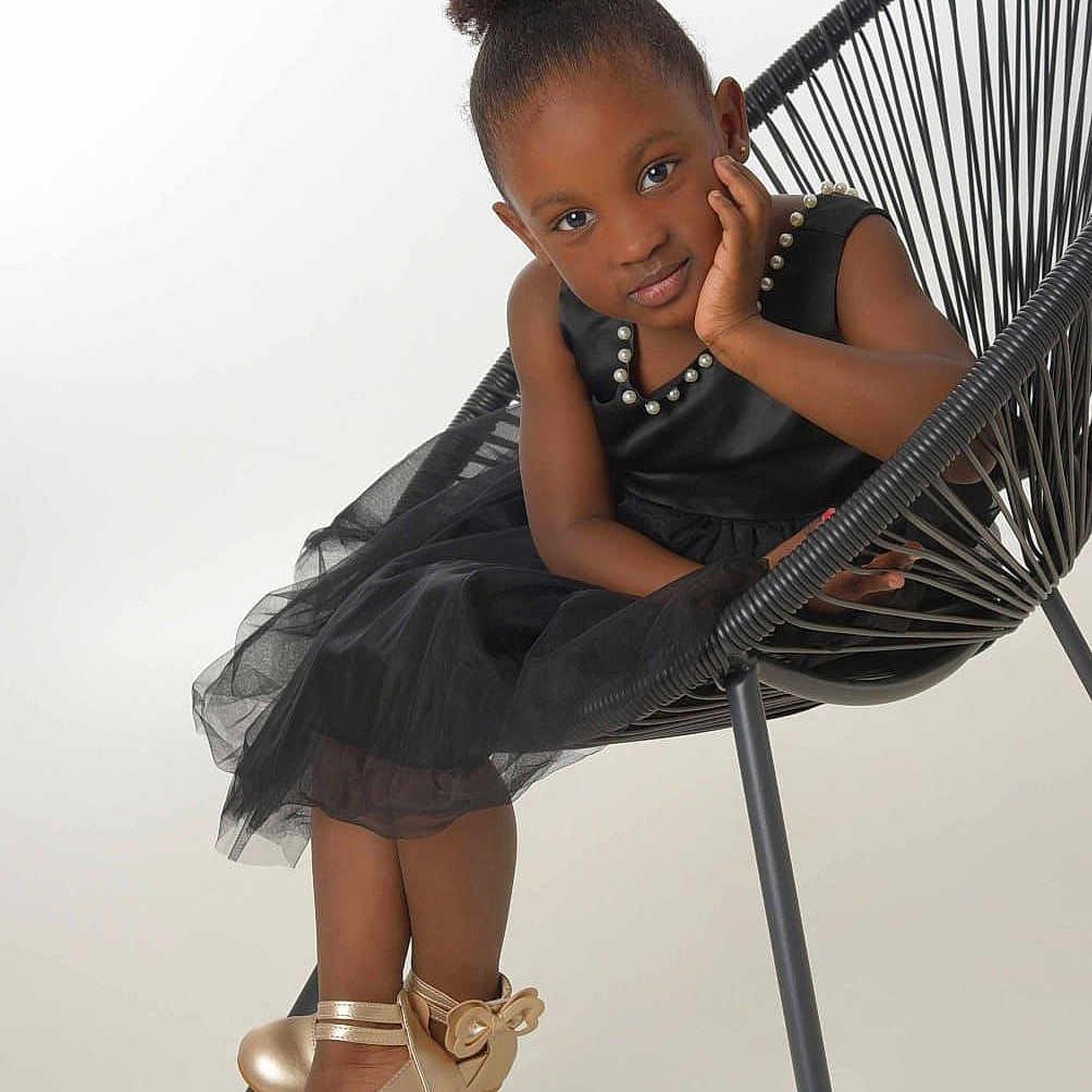 Binta a rejoint le concours — aidez-le/la à gagner de superbes lots ! black_dress, child, curly_hair, cute, elegant, expression, fashion, footwear, girl, gold_shoes, indoor, modern_chair, pearl_accessory, portrait, pose, sitting, smiling, studio, white_background, young