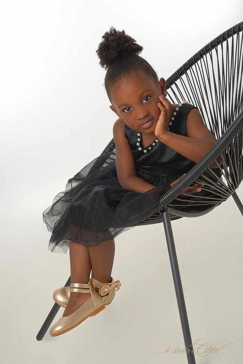 Binta a rejoint le concours — aidez-le/la à gagner de superbes lots ! child, girl, black_dress, gold_shoes, modern_chair, sitting, portrait, studio, fashion, cute, pearl_accessory, smiling, curly_hair, elegant, young, pose, footwear, white_background, expression, indoor