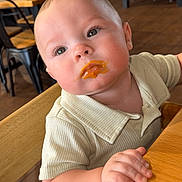 Russell is registered to the contest to win money with this photo: baby, child, high_chair, food, face, curious, indoor, wooden_furniture, table, restaurant, orange_food, clothing, short_sleeve, person, cute, portrait, seat, cafe, plush_cheeks, dining