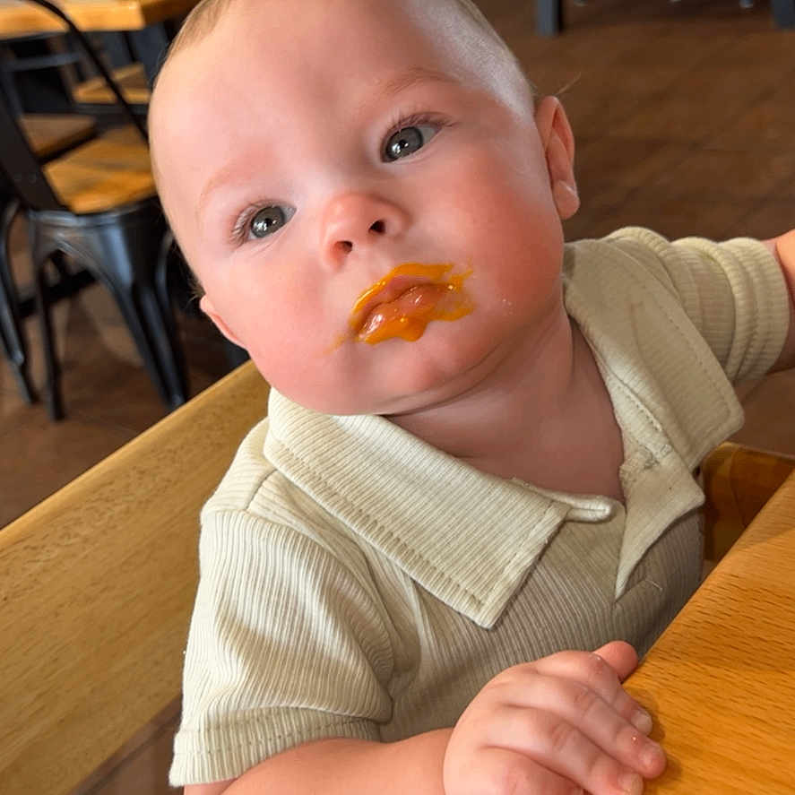 Russell is registered to the contest to win money with this photo: baby, child, high_chair, food, face, curious, indoor, wooden_furniture, table, restaurant, orange_food, clothing, short_sleeve, person, cute, portrait, seat, cafe, plush_cheeks, dining