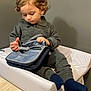 baby, bag, changing_pad, child, curious, curly_hair, exploring, floor, grey_clothing, home, indoor, neutral_wall, person, sitting, socks, table, toddler, toy, washing_machine, young_child
