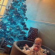 Lena is registered to the contest to win money with this photo: baby, ball_pit, balls, child, christmas_decorations, christmas_tree, cozy, cute, festive, gift_wrap, happy, holiday, indoors, lighting, presents, sitting, smiling, toy, wall, wooden_floor