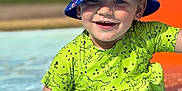 Lyam a rejoint le concours — aidez-le/la à gagner de superbes lots ! child, toddler, smiling, pool, water, splashing, hat, bucket_hat, blue, green_shirt, dinosaur_print, sunlight, outdoor, happy, summer, play, cute, face, person, fun