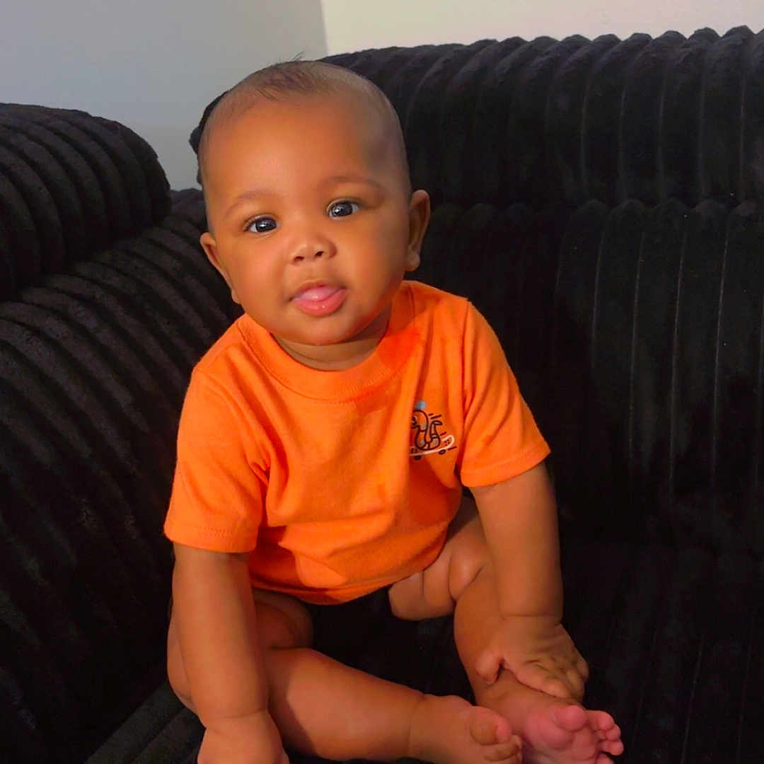 Aahshon is registered to the contest to win money with this photo: baby, black_couch, casual_clothing, child, cute, eyes, face, foot, hand, head, home, indoor, infant, orange_shirt, person, plush_furniture, sitting, skin, smiling, tongue_out