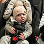 baby, car_seat, child, closeup, comfort, cushion, cute, gray_clothing, headwear, indoor, infant, knit_hat, patterned_fabric, person, playful, pom_pom, seat_belt, sitting, soft_lighting, tongue_out