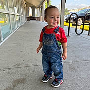 Lorenzo is registered to the contest to win money with this photo: bracelet, building, car, casual, child, curious, daylight, denim_overalls, glass_windows, outdoor, parking_lot, person, red_shirt, sidewalk, smiling, sneakers, standing, tattoo, toddler, urban