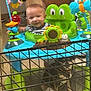Adan is registered to the contest to win money with this photo: baby, child, activity_center, frog_design, toy, dog, cage, indoor, smiling, face, animal, pet, playtime, colorful, green, blue, orange, plastic, floor, person
