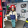 Lucas participe au concours pour gagner de l'argent avec cette photo : bag, blue, cabinet, casual_clothing, child, childrens_fashion, emergency_vehicle, firefighter_helmet, floor, helmet, indoor, jacket, medical_equipment, oxygen_tanks, pants, red, smiling, sneakers, standing, white