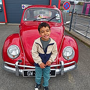 Lucas participe au concours pour gagner de l'argent avec cette photo : asphalt, boy, casual_clothing, child, daylight, fence, happy, jacket, license_plate, outdoor, parking_lot, person, red_car, road_sign, sign, smile, sneakers, standing, vehicle, vintage_car