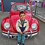 asphalt, boy, casual_clothing, child, daylight, fence, happy, jacket, license_plate, outdoor, parking_lot, person, red_car, road_sign, sign, smile, sneakers, standing, vehicle, vintage_car