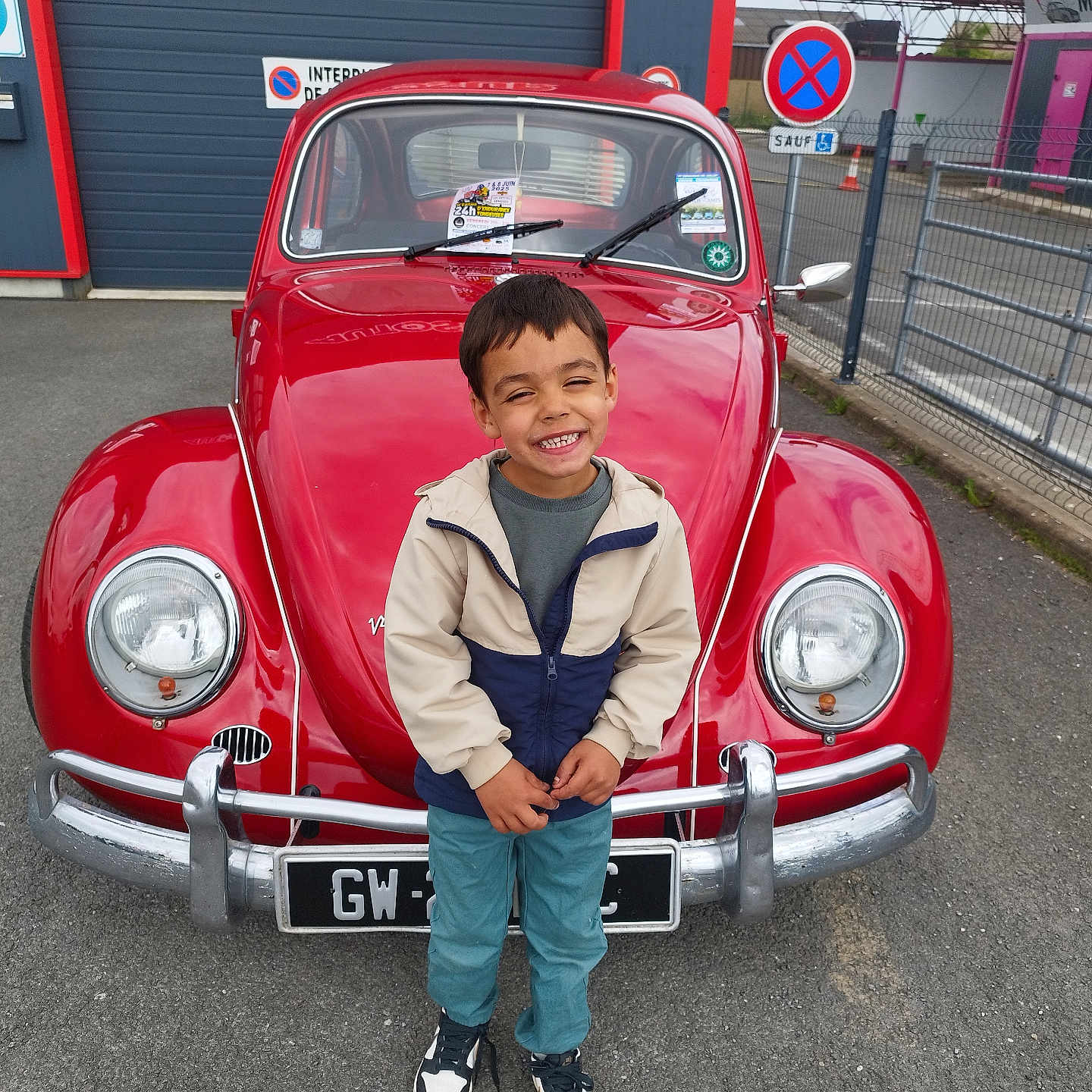 Lucas participe au concours pour gagner de l'argent avec cette photo : asphalt, boy, casual_clothing, child, daylight, fence, happy, jacket, license_plate, outdoor, parking_lot, person, red_car, road_sign, sign, smile, sneakers, standing, vehicle, vintage_car