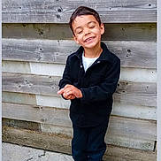 Lucas a rejoint le concours — aidez-le/la à gagner de superbes lots ! child, boy, smiling, closed_eyes, black_jacket, white_tshirt, dark_pants, sneakers, wooden_wall, outdoor, concrete_floor, standing, happy, casual_clothing, portrait, person, fashion, playful, young, cute
