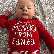 Jagger is registered to the contest to win money with this photo: baby, infant, onesie, red_clothing, striped_waistband, floral_bed_sheet, bed, lying_down, face, eyes_open, gaze, cute, newborn_look, portrait, holiday_theme, christmas_text, white_text, cozy, indoor, human