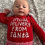 baby, infant, onesie, red_clothing, striped_waistband, floral_bed_sheet, bed, lying_down, face, eyes_open, gaze, cute, newborn_look, portrait, holiday_theme, christmas_text, white_text, cozy, indoor, human