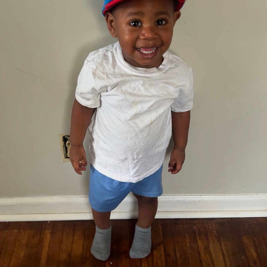 Makai is registered to the contest to win money with this photo: blue_shorts, casual_clothing, child, cute, feet, happy, hat, headwear, indoor, mickey_mouse, person, portrait, smiling, socks, standing, toddler, wall, white_tshirt, wooden_floor, young_child
