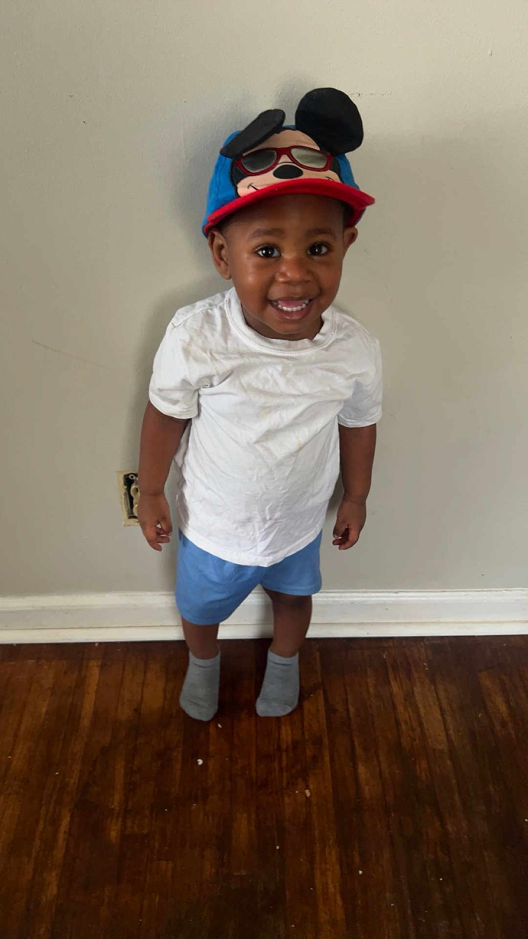Makai is registered to the contest to win money with this photo: toddler, child, smiling, hat, mickey_mouse, white_tshirt, blue_shorts, socks, standing, indoor, wooden_floor, wall, happy, casual_clothing, person, portrait, cute, young_child, headwear, feet