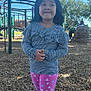 child, playground, outdoor, trees, wood_chips, play_structure, clothing, striped_shirt, leggings, hearts, shoes, person, smile, standing, daylight, fun, park, young, casual_wear, expression