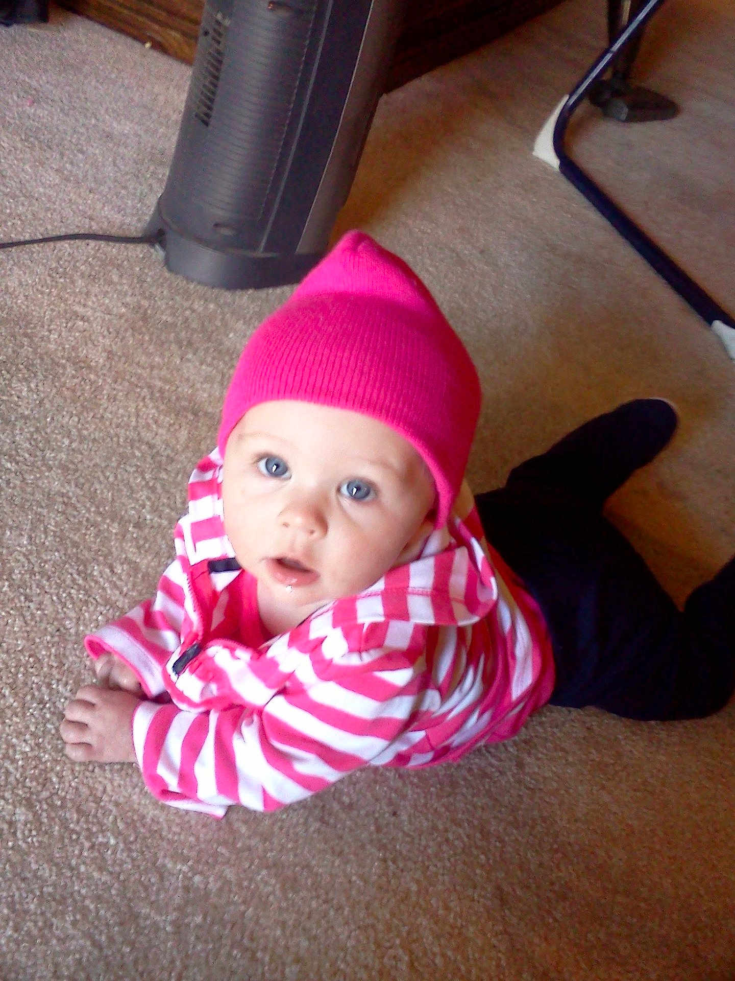 Katelynn is registered to the contest to win money with this photo: baby, child, pink_hat, striped_jacket, carpet, indoor, heater, floor, person, blue_eyes, clothing, crawling, cute, young_child, infant, headwear, homedecor, furniture, warm_clothing, expression