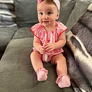 Sapphire is registered to the contest to win money with this photo: baby, child, pink_bow, headband, pink_outfit, socks, brush, couch, gray_couch, blanket, indoor, smiling, cute, infant, seated, person, portrait, cozy, soft_lighting, home