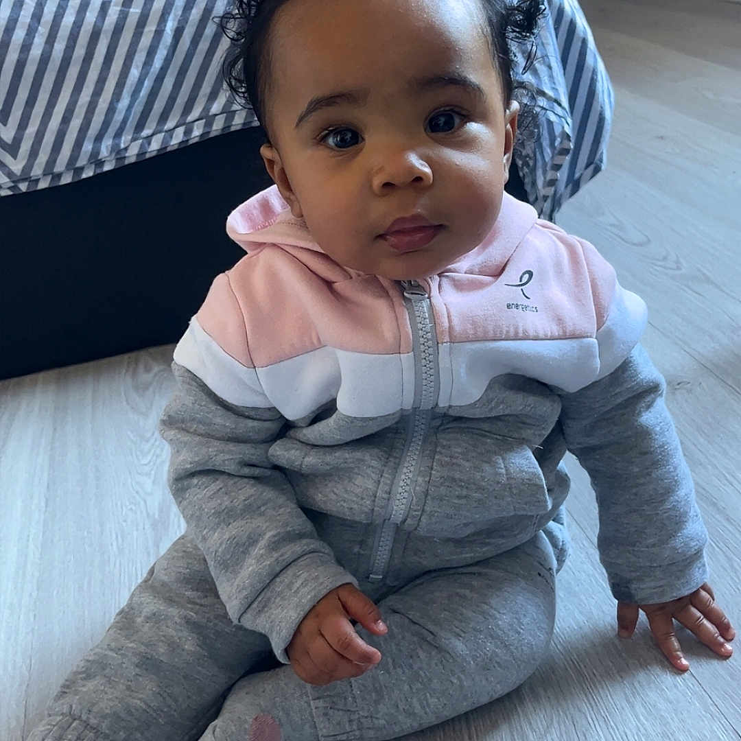 Anaya a rejoint le concours — aidez-le/la à gagner de superbes lots ! baby, blanket, casual_wear, child, curly_hair, cute, expression, face, floor, grey_clothing, hoodie, indoor, person, pink_clothing, portrait, sitting, striped_bed, toddler, wooden_floor, young_child