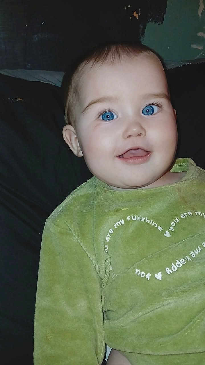 Hayzleigh joined the competition — help win amazing prizes! baby, infant, blue_eyes, smile, face, portrait, green_clothing, onesie, close_up, indoor, pillow, bed, dark_background, chubby_cheeks, ear, skin, happy, camera_flash, cute, sitting