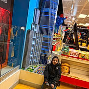 Manuel is registered to the contest to win money with this photo: child, black_jacket, lego_store, lego_buildings, lego_cityscape, lego_boxes, toy_store, floor_tiles, yellow_floor, game_boy_lego, sitting, indoor, display_shelves, shopping, store_lighting, lego_characters, people, shopping_cart, retail_display, casual_clothing