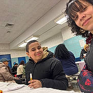 Manuel joined the competition — help win amazing prizes! backpack, bench, boy, casual_clothing, child, community, conversation, curly_hair, event, fluorescent_lighting, indoor, jacket, marker, paper_plate, people, seated, smile, table, wall_tiles, woman